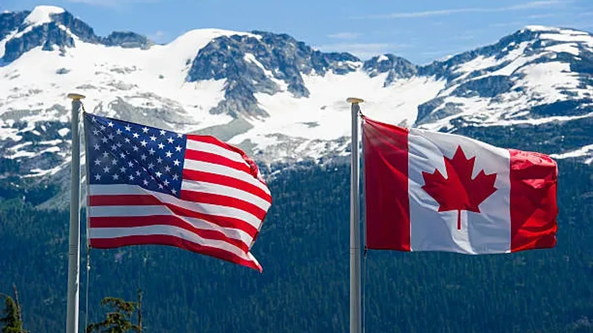 USA and Canada flags wave in the wind with mountains in the background.