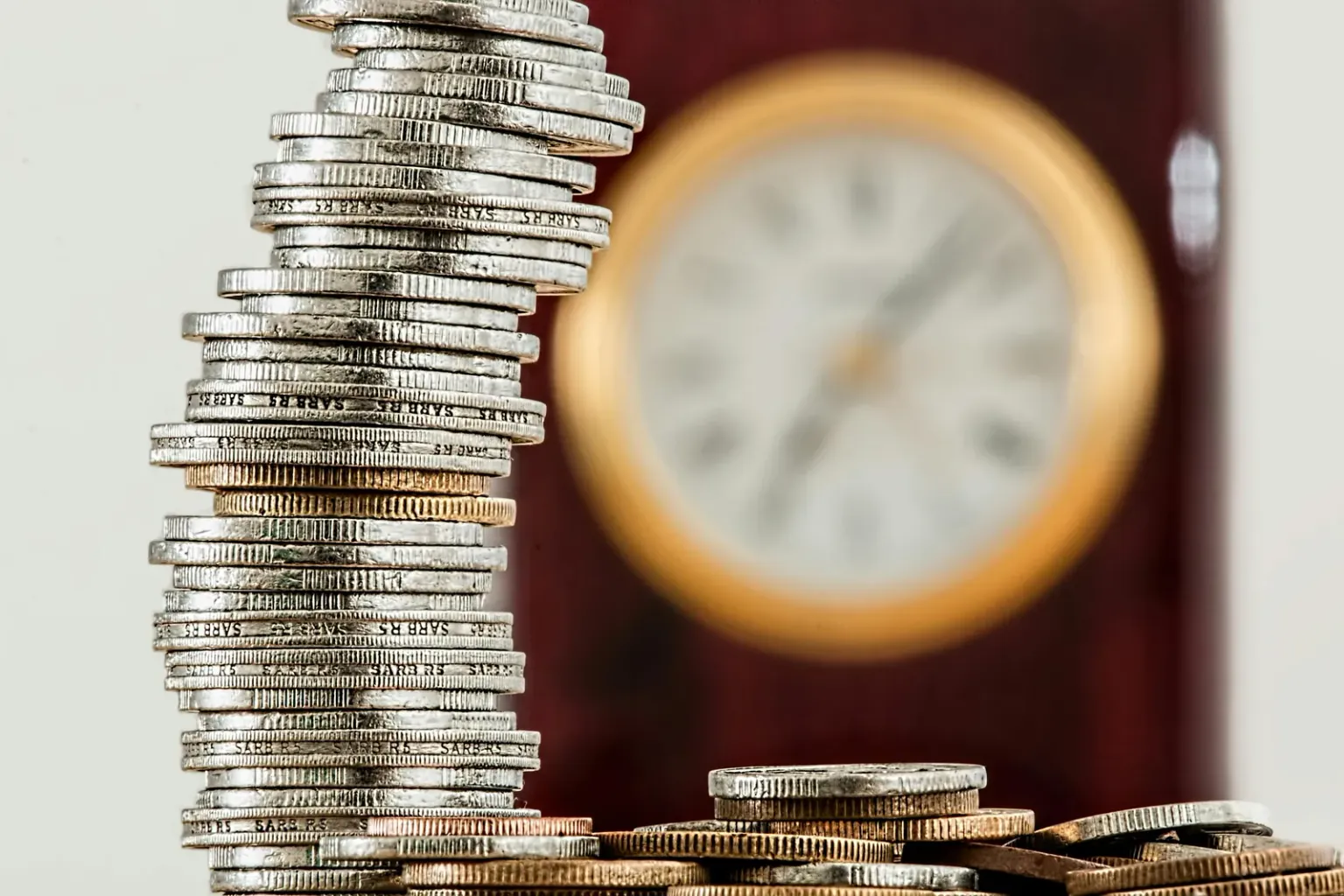 Gros plan d'une pile de pièces de monnaie avec quelques pièces éparpillées à la base. En arrière-plan, une horloge floue donne une impression de temps qui passe.