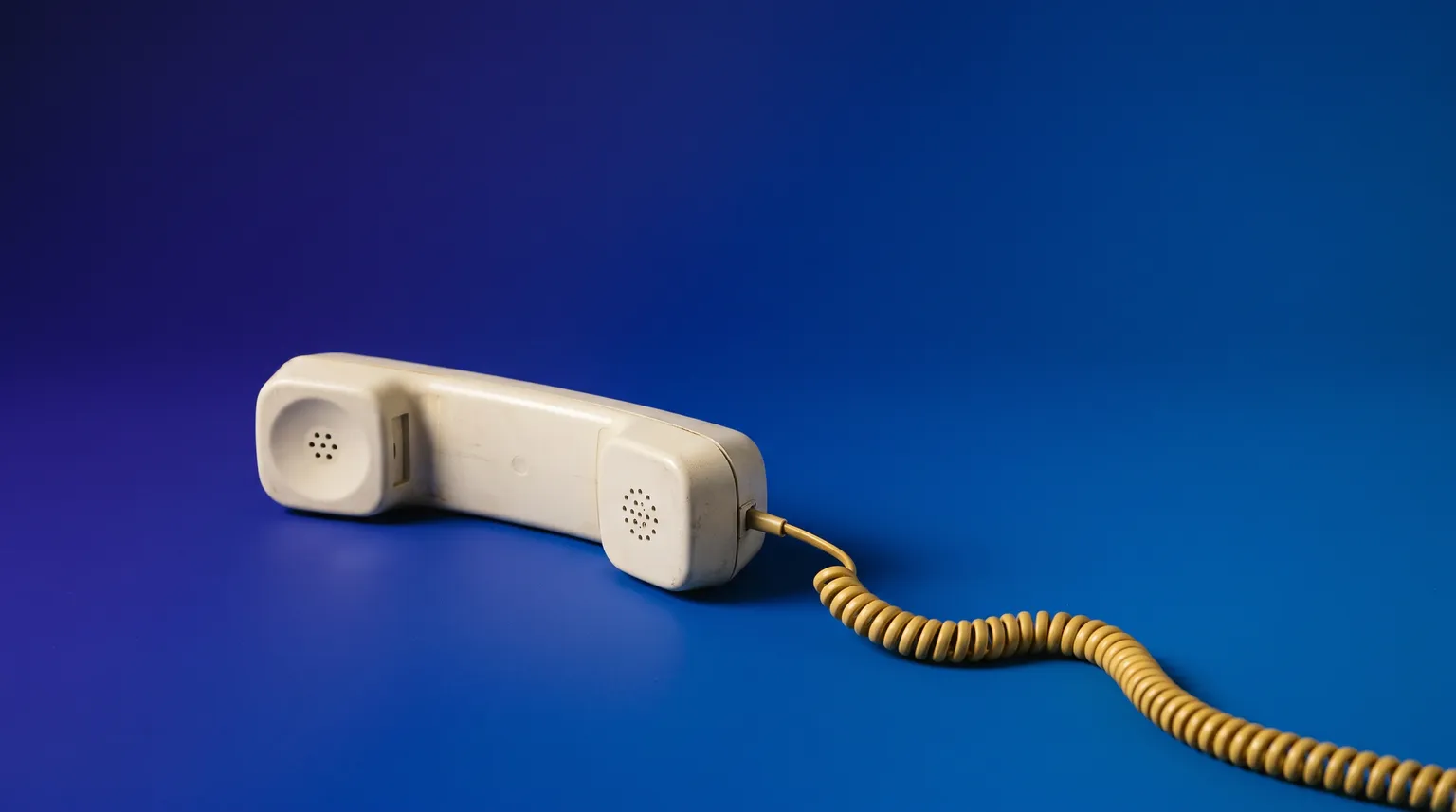 An off-white corded telephone handset lying off the hook on a deep blue backdrop, its coiled cord in gold.