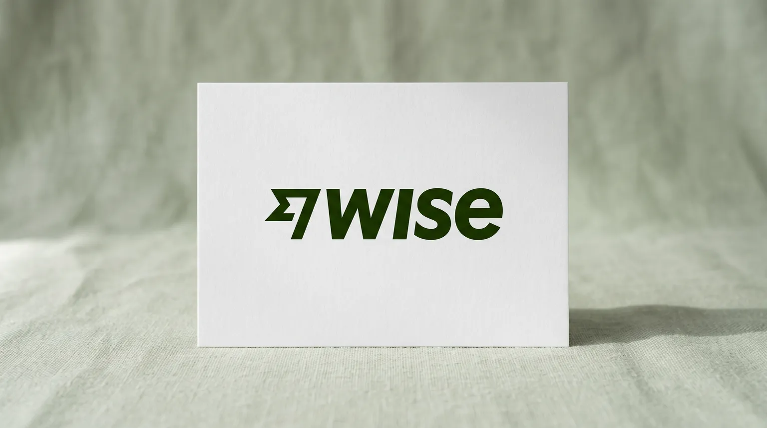 The Wise logo on a matte white card standing on a pale sage-green linen surface in soft window light.
