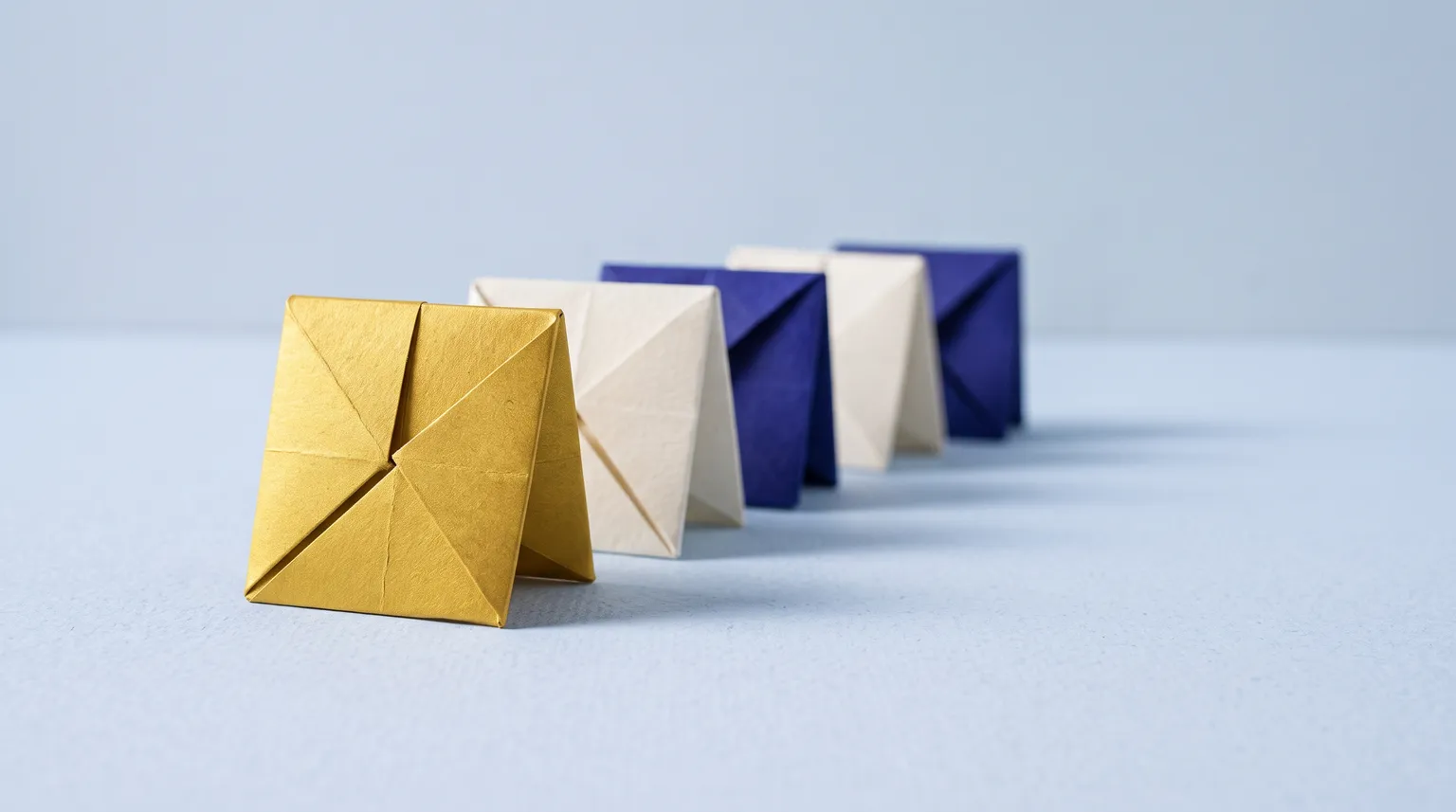 Five folded paper tiles in a deliberate row on a pale blue ground, the first one gold.